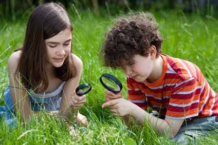 Two children using magnifying glasses to explore grass in a park, showcasing curiosity and outdoor learning.